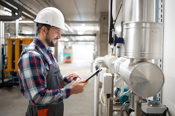 Installateur im Heizungraum steht an den Leitungen und prüft mit dem Tablet die Werte