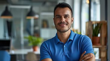 Porträtbild junger Mann mit dunklem Dreitagebart und blauem Poloshirt, im Hintergrund Wohnung