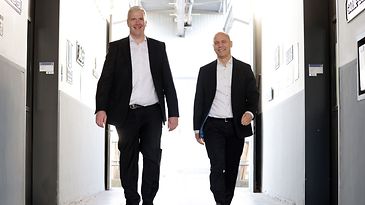 Zwei Männer in dunklen Anzügen gehen Flur entlang, auf Kamera zu